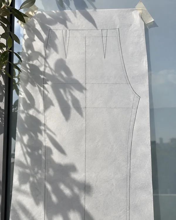 A trouser pattern draft in fine pencil on translucent tracing paper, taped to a glass panel in sunlight.