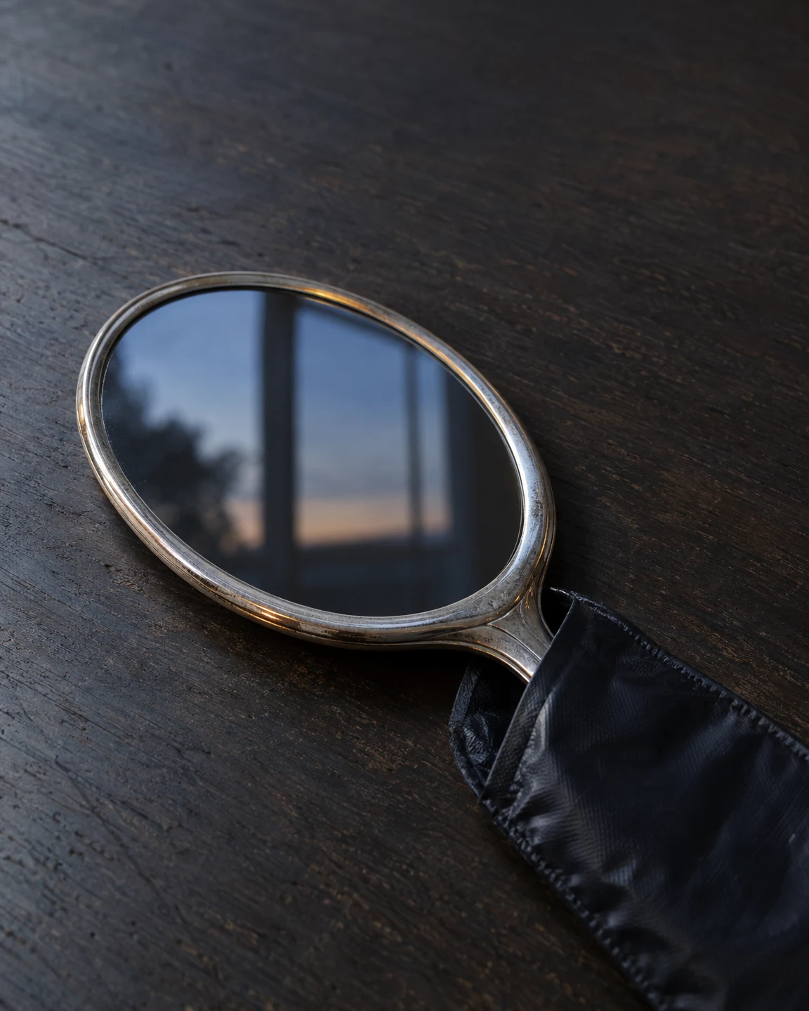 A vintage oval silver hand mirror lies diagonally on a charcoal wooden tabletop, reflecting a blue window and dark outdoor silhouettes; its handle extends into…