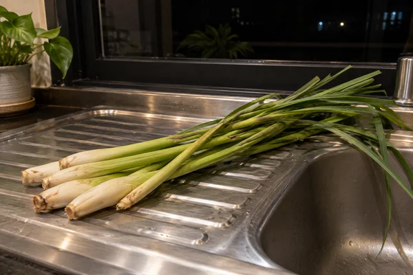Several pale-based lemongrass stalks lie diagonally across a ribbed stainless-steel sink draining board, with long green leaves extending over the basin.