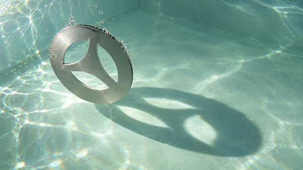 A circular brushed-metal disc with three leaf-shaped openings hangs by a thin line in pale green water, casting a broad shadow and bright patches across the…