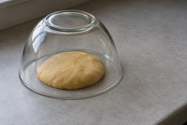 A thick oval of golden dough rests beneath an inverted clear glass bowl on a grey counter, with open counter space extending to the right and a window…