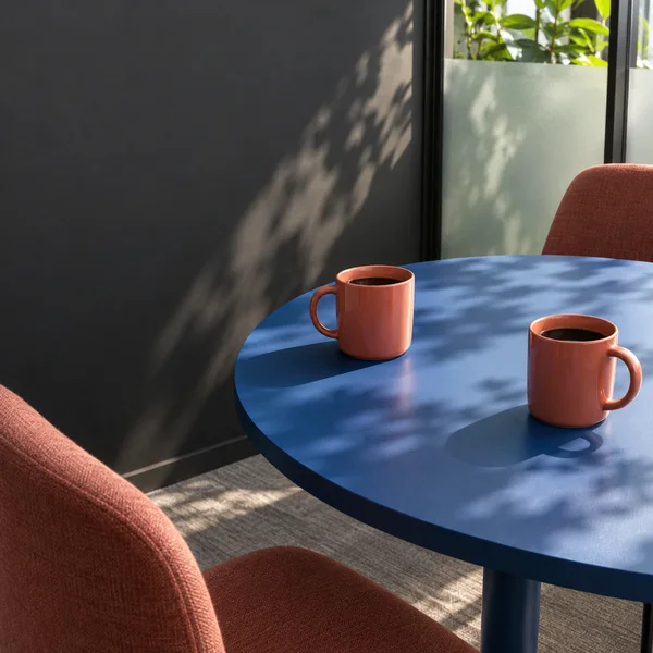 A cobalt-blue round table holds two coral mugs in a sunlit office corner, with cropped coral-upholstered chairs, a frosted-glass partition, and foliage visible…