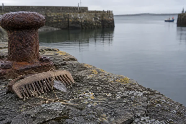 A weathered wooden comb with pale grass strands and a grey feather rests on a lichen-covered stone harbour quay beside a large rusted mooring bollard, with…