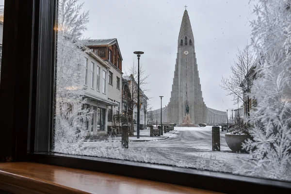View through a frosted café window toward Hallgrímskirkja in Reykjavík, with its clock and stepped concrete wings above a lightly snow-covered street.