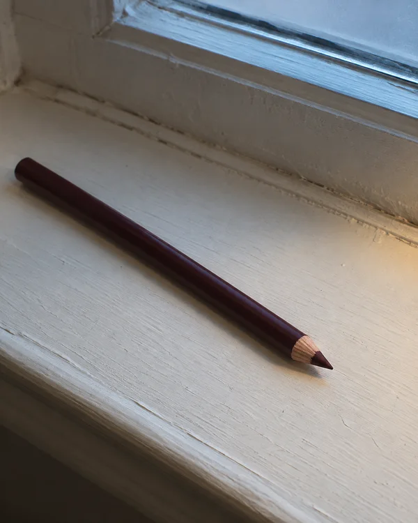 A sharpened burgundy pencil lies diagonally across a pale, weathered painted windowsill beneath a blue-lit window, with its exposed wood and dark point visible.