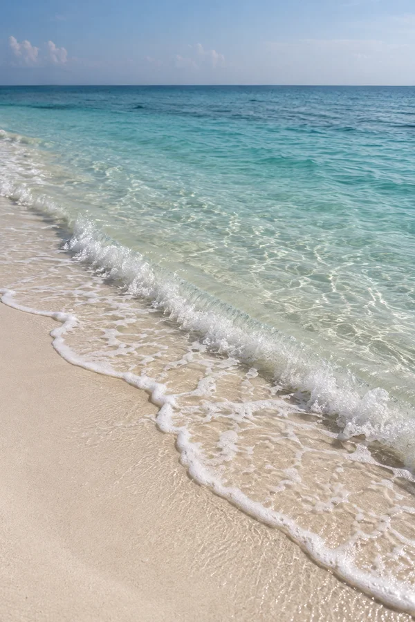 A small foamy wave breaks diagonally across pale coral-colored sand, with clear sea-green shallows revealing the sandy bottom and muted blue water beneath a…