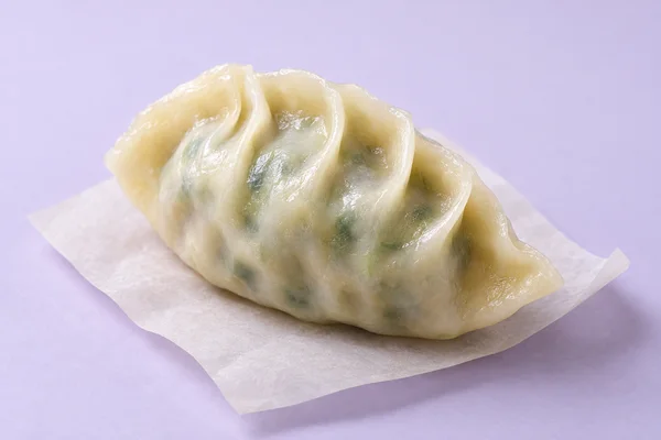 A glossy, pale crescent dumpling with visible green filling and curved pleats rests on a sheet of ivory paper against a lavender background.