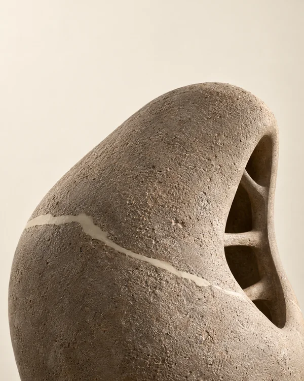 A close-up of a softly rounded, rough taupe sculpture against a warm ivory background, with a pale repair line across its front and curved ribs visible through…