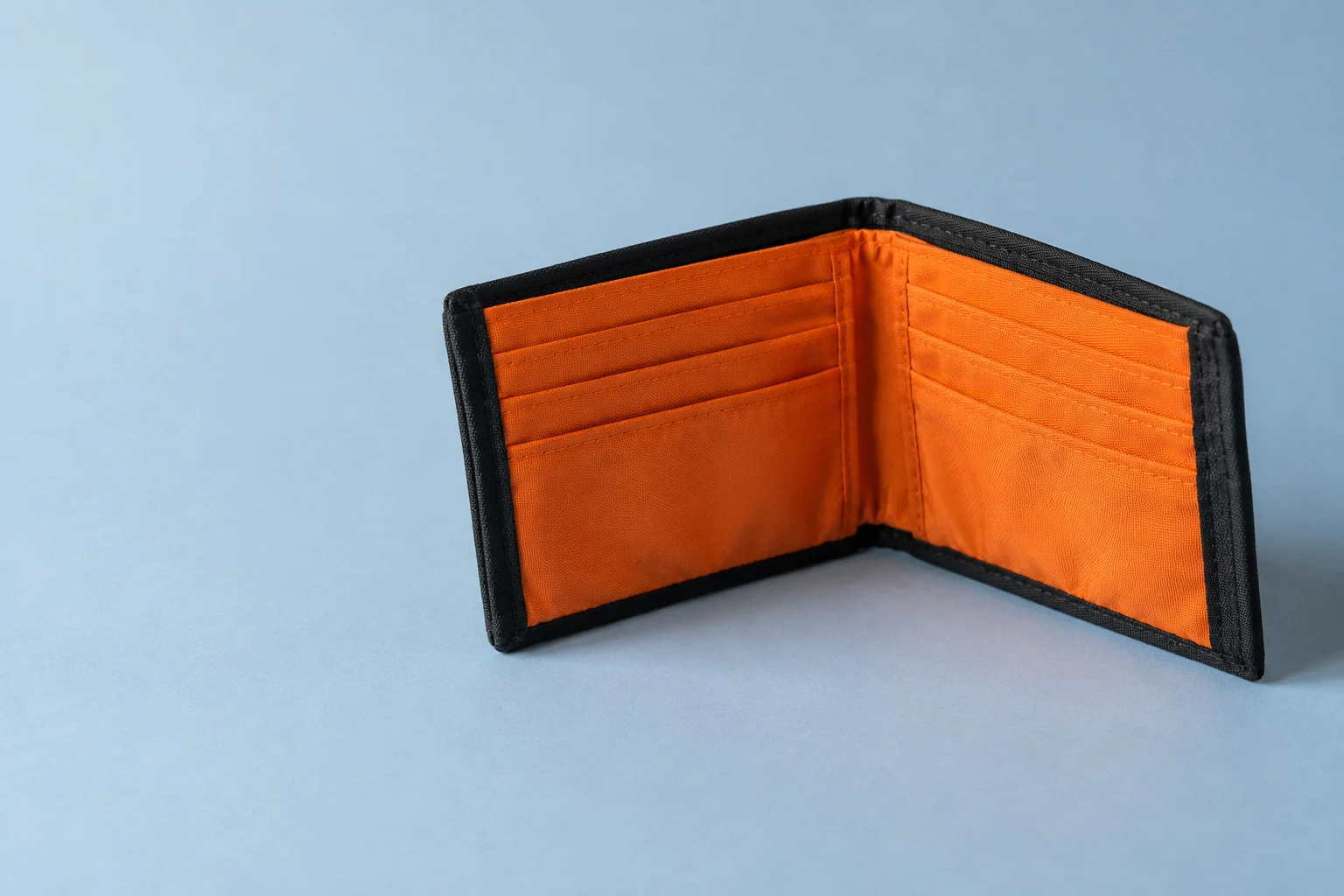 An open black fabric bifold wallet with vivid orange lining and empty card pockets sits on a pale blue surface, with ample blank space to the left.