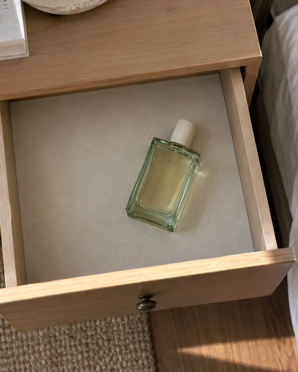 A pale-green glass fragrance bottle with a plain ivory cap lies diagonally in a cream-lined maple dresser drawer, viewed from above; the open drawer sits…