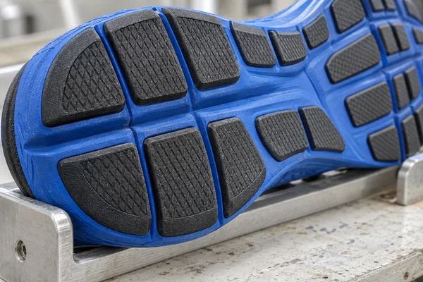 Close-up of a blue trainer outsole with charcoal tread blocks resting in a brushed-aluminum support on a lightly scuffed workbench.