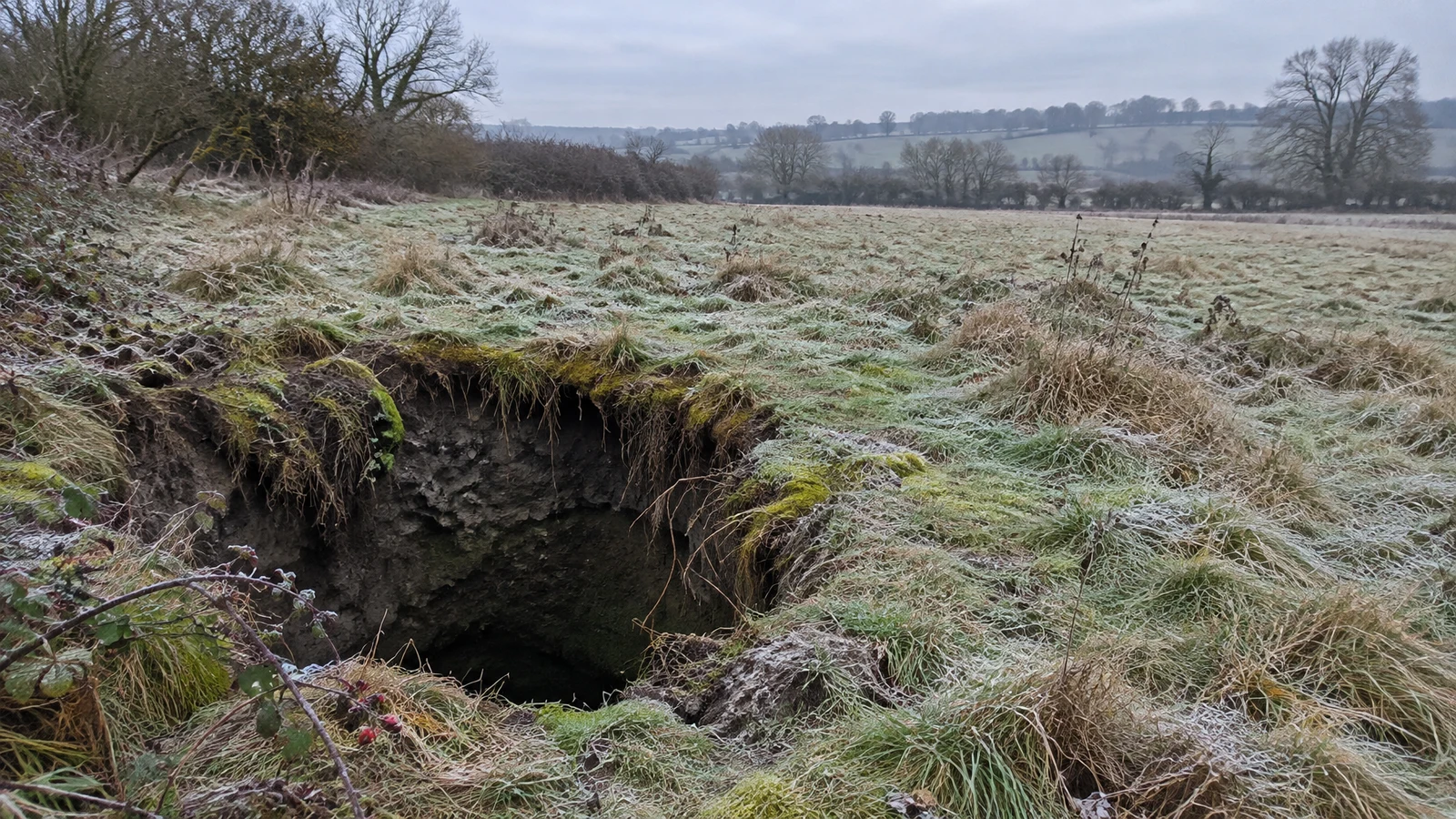 A large dark sinkhole opens in a frost-dusted meadow, its uneven earthen rim covered with grass, exposed roots and moss.
