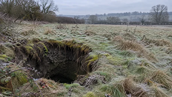 A large dark sinkhole opens in a frost-dusted meadow, its uneven earthen rim covered with grass, exposed roots and moss.