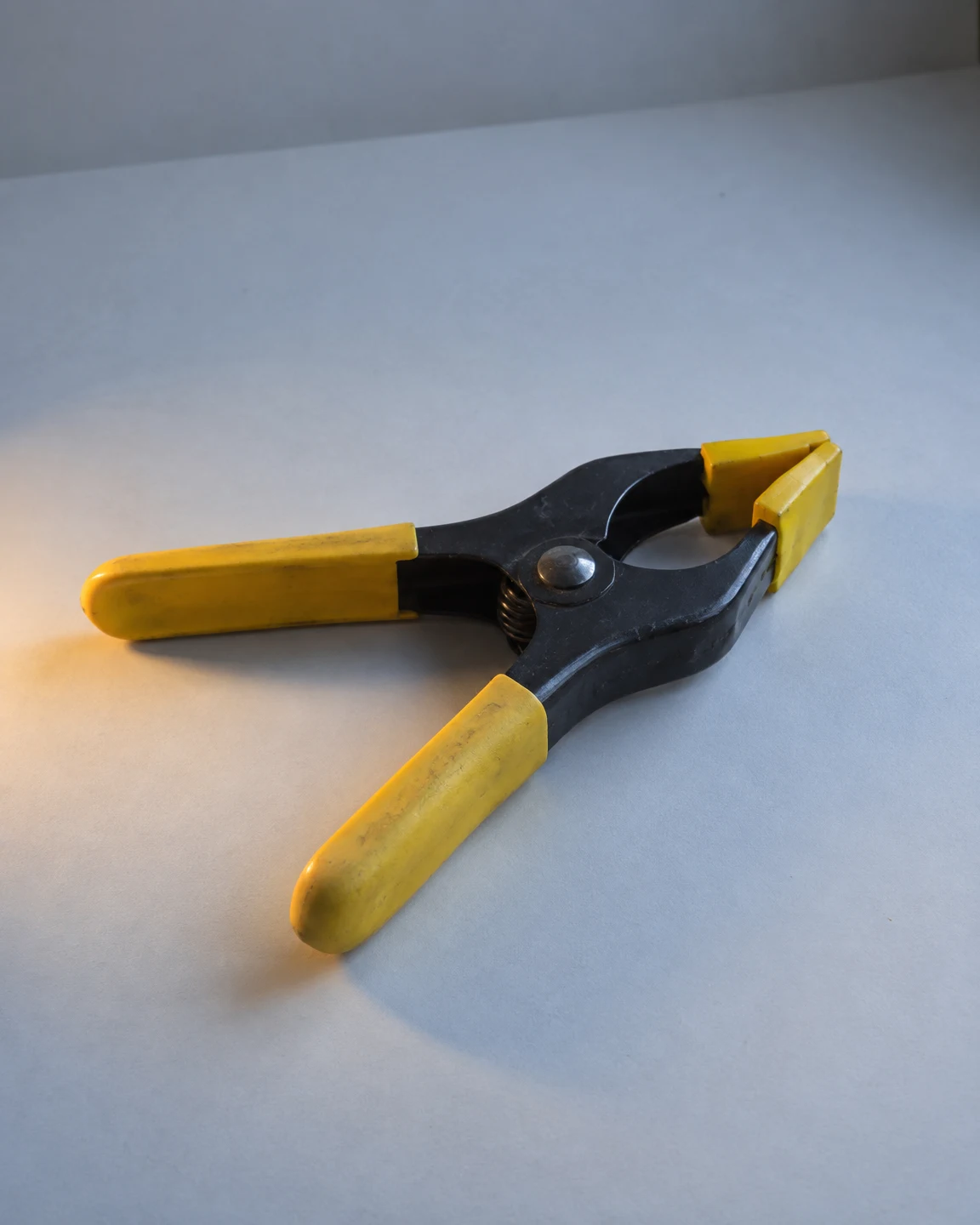 A black steel spring clamp with yellow rubber sleeves and jaw pads lies diagonally on a pale gray studio sweep, lit by cool blue-gray light and a warm amber…
