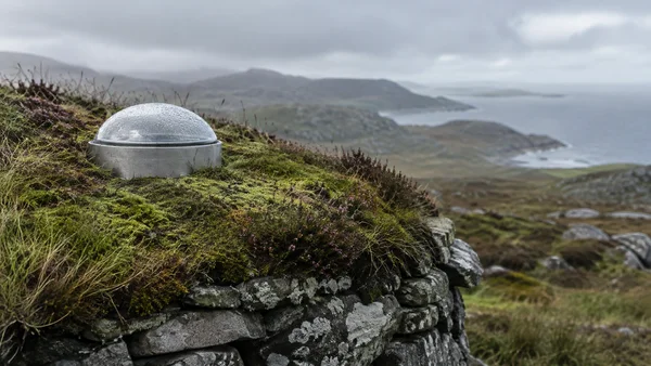 A mist-beaded clear dome on a brushed-metal base sits in the moss and heather covering a stone roof, with a hazy coastal hillside and sea behind it.