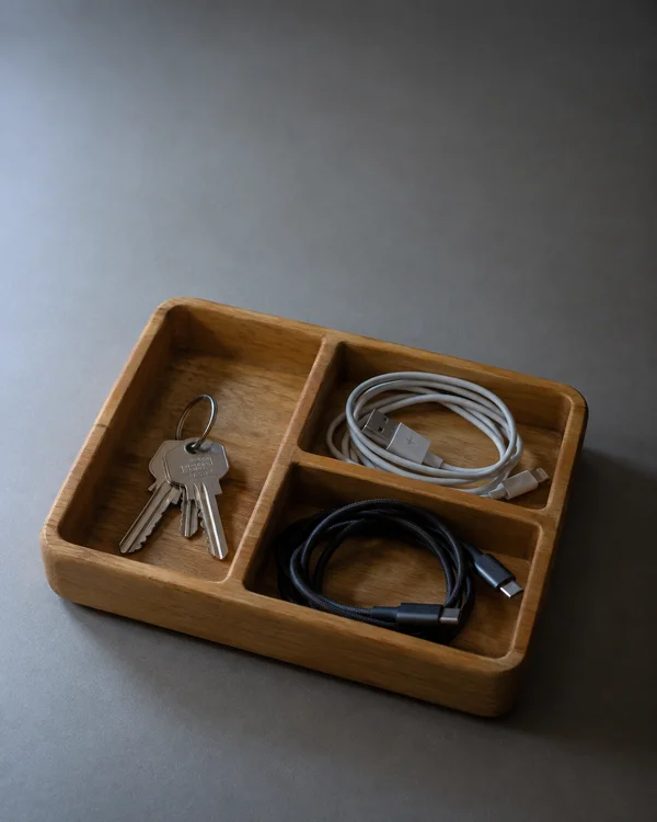 A honey-coloured wooden organiser tray holds a metal key ring in one compartment and a white cable and a black cable in the other two, against a warm-grey…