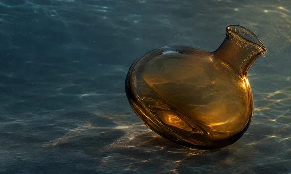 A broad, amber glass carafe lies on its side on a dark slate-blue surface, its flared pouring lip angled toward the upper right.