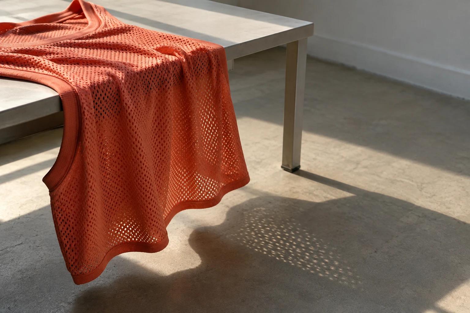 A coral mesh basketball jersey drapes over the near edge of a pale grey table, casting a perforated shadow across the warm grey floor in direct sunlight.