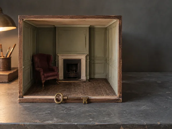 A handmade open-front miniature parlour sits on a slate-grey worktable, with sage panelled walls, a cream fireplace, a burgundy wingback chair and a large…