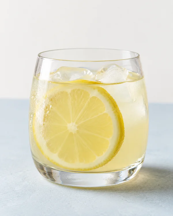 A clear rounded tumbler of pale-yellow lemonade with two visible ice cubes and a large lemon wheel inside, set on a pale blue tabletop against an off-white…
