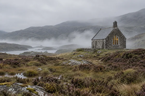 A small weathered stone building with a slate roof, bell tower and glowing arched window stands on a misty moor above a quiet loch, surrounded by wet heather…
