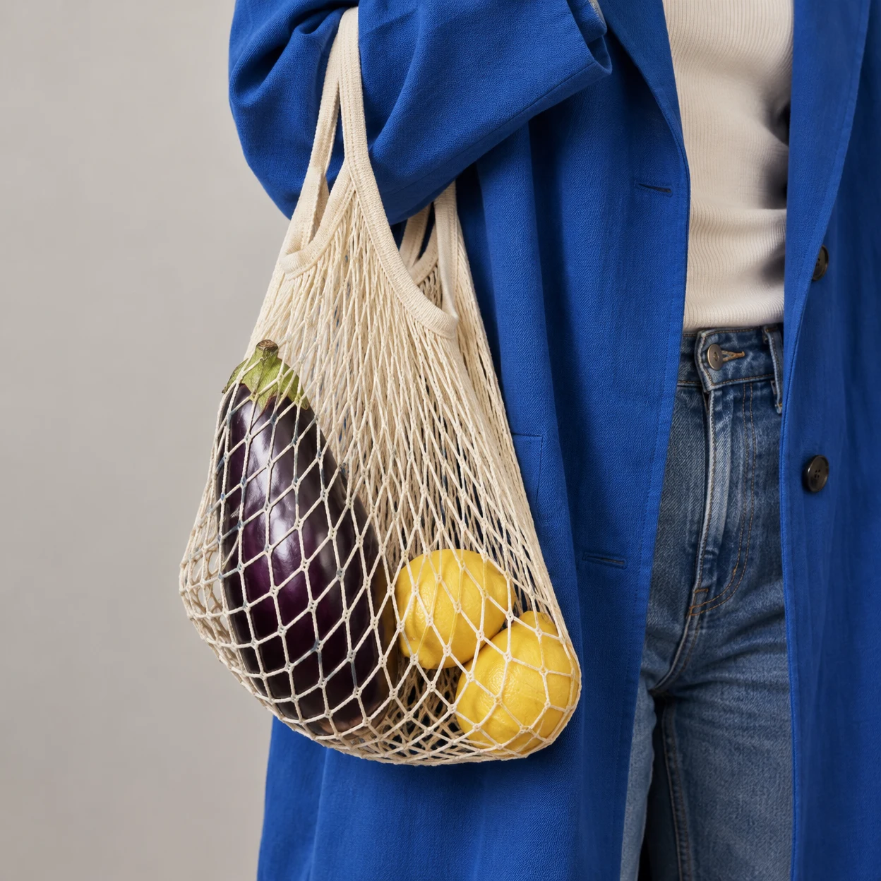 A cream mesh grocery bag containing one purple aubergine and two yellow lemons hangs beside a cobalt-blue coat against a pale warm-grey wall; the cropped…