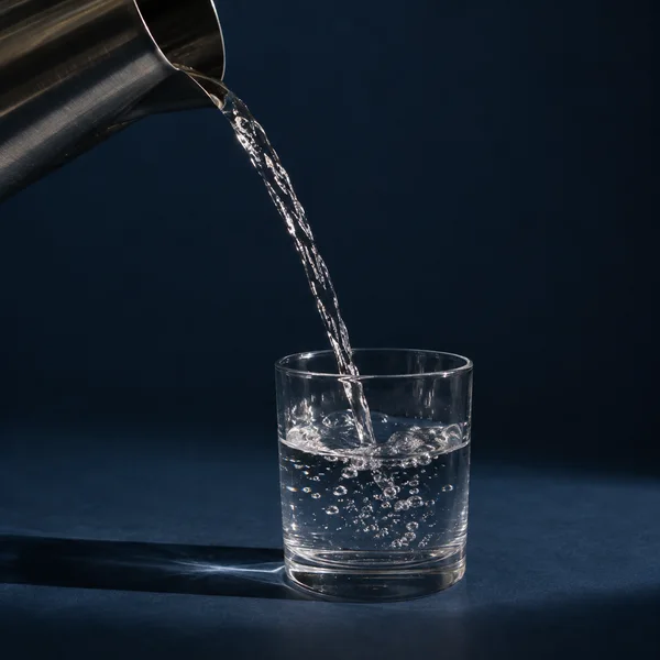 A stainless-steel pitcher pours a bright stream of water into a clear tumbler on a dark blue surface, with bubbles in the glass and a long shadow extending to…