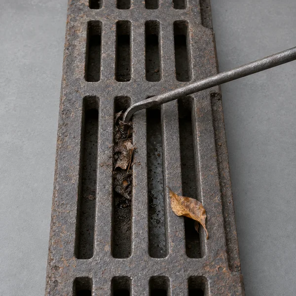 A weathered cast-iron storm-drain grate lies vertically on a cool-gray surface, with damp leaves and silt packed into one slot; a steel hook reaches in from…