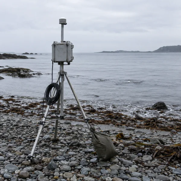 A weathered metal tripod with a grey field instrument box and short mast stands on a pebble beach beside a calm sea; a coiled black cable hangs from the rig…