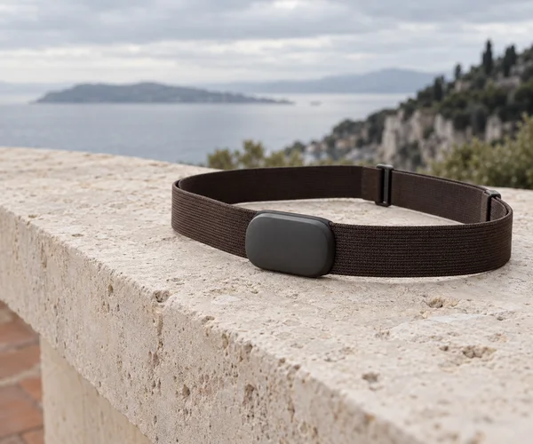 A charcoal sensor module attached to a dark brown woven chest strap rests in an oval on a pale stone ledge, with a softly blurred coastal view under an…