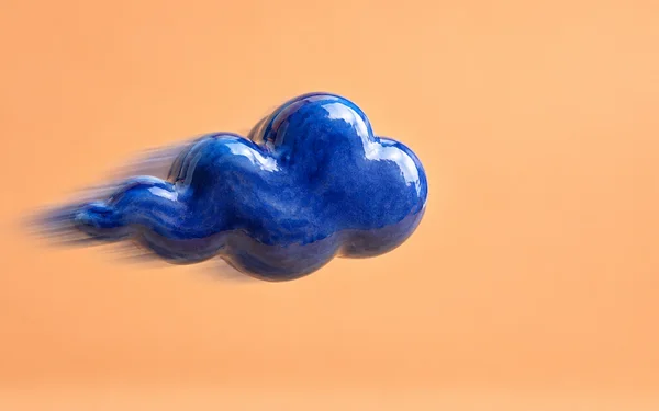 A glossy cobalt-blue ceramic cloud with rounded lobes and a tapered left tail appears to fly across a warm apricot background, with motion streaks behind it.