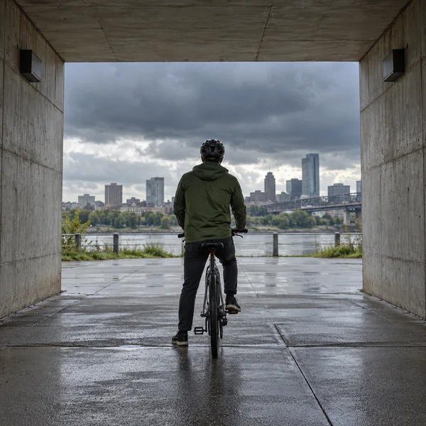 A helmeted cyclist in a muted green jacket pauses astride a bicycle beneath a concrete underpass, facing a wet pavement, river, and city skyline beneath dark…
