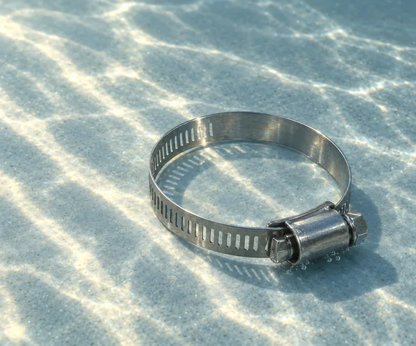 A stainless-steel worm-drive hose clamp rests in a circular loop on a pale blue-grey submerged floor, with its screw housing at the lower right, small bubbles…