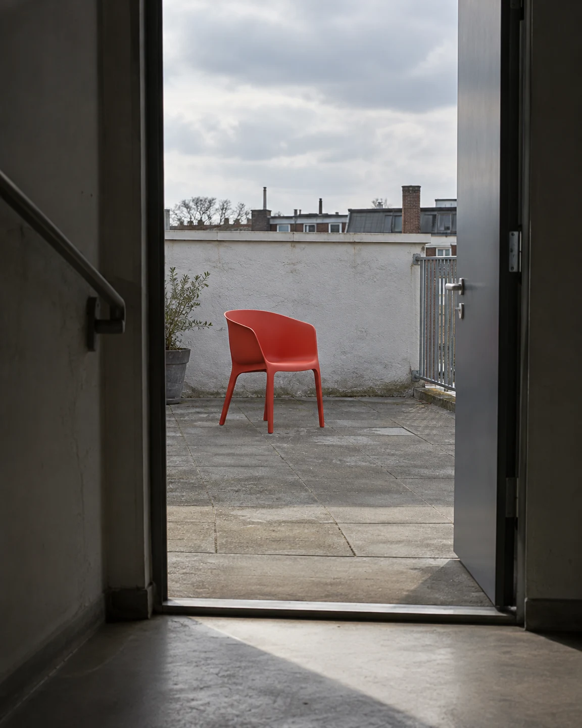 A coral-red molded chair sits on a paved rooftop terrace, viewed through an open doorway from a dim stairwell.