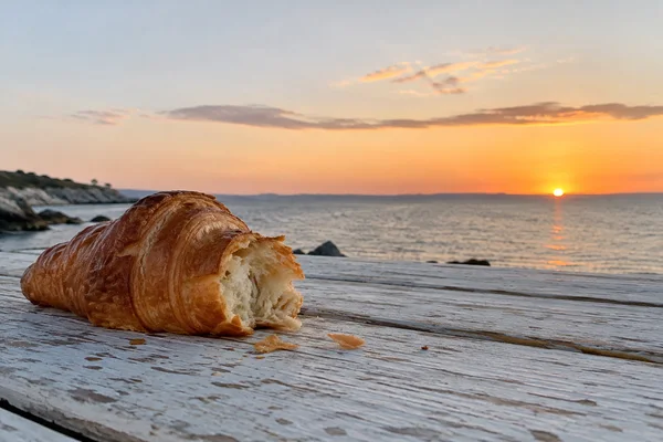 A large torn croissant with scattered flakes rests on a pale weathered wooden table, with the sea and a peach-gold sunset behind it.