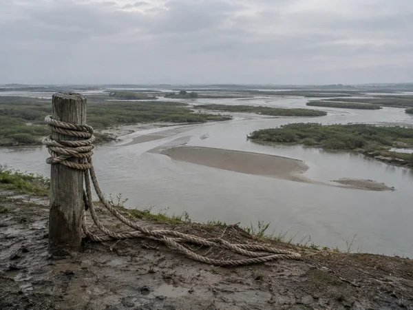 A weathered wooden post wrapped in thick rope stands on a muddy riverbank, with rope lengths lying across the foreground.