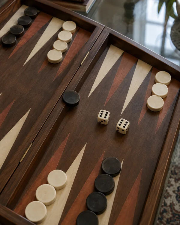 An overhead oblique view of a wooden backgammon board on a glass table, with two ivory dice showing sixes near the center, a dark checker on the bar, and ivory…