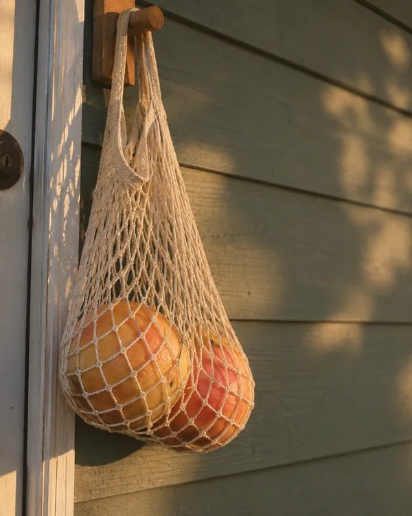 A cream cotton net bag containing two yellow-pink grapefruits hangs from a wooden peg beside a door, against muted green porch siding in warm low-angle…