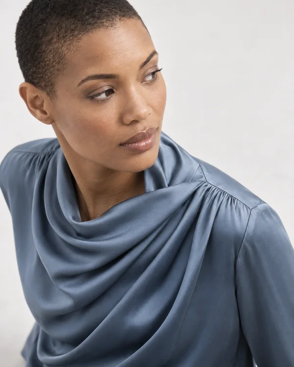 A close studio portrait of a short-haired model in a blue-gray draped blouse with gathered shoulder seams, looking off-frame against a pale seamless background.