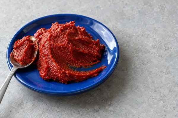 A large mound of deep red tomato paste with a curved smear sits on a glossy cobalt-blue saucer on a pale-grey stone tabletop.