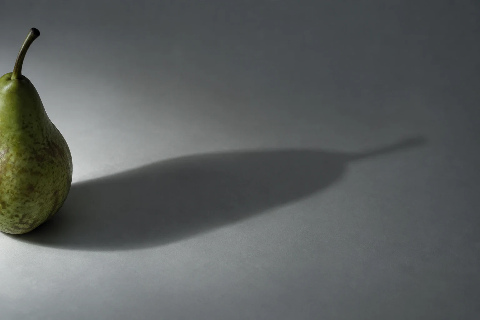 A mottled green pear is partly cut off at the left edge of a grey surface, casting a long shadow diagonally across the open space to the right.