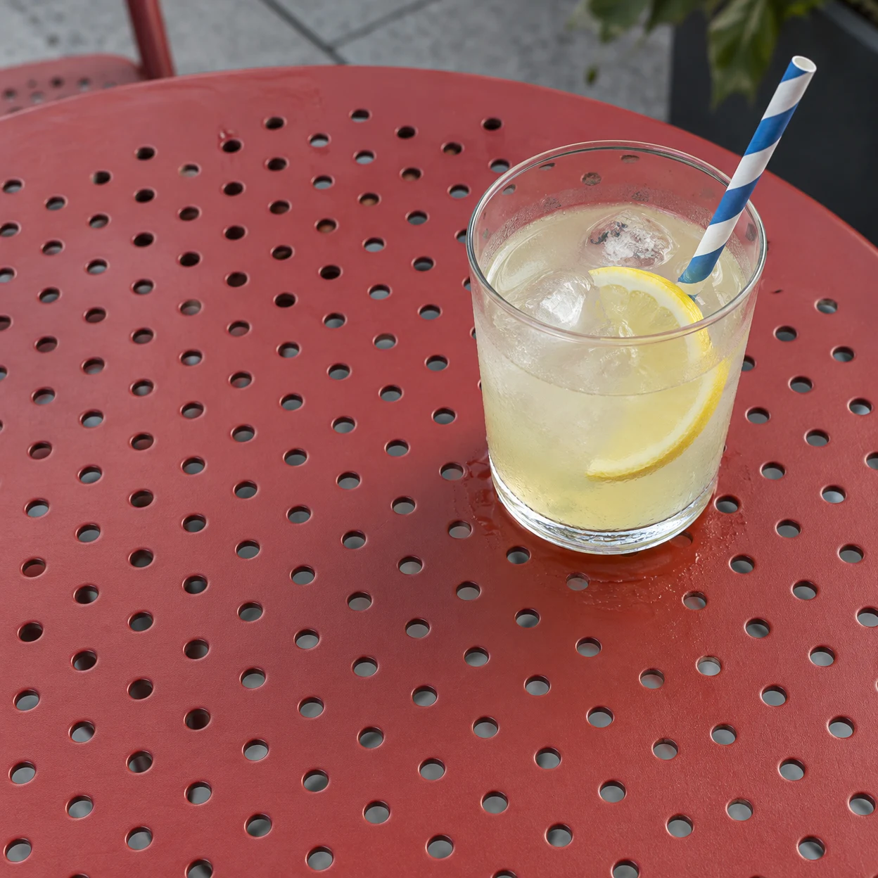 A clear tumbler of pale yellow lemonade with ice and a lemon slice sits on a red perforated metal café table.