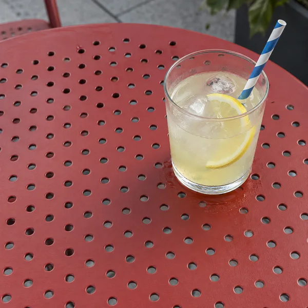 A clear tumbler of pale yellow lemonade with ice and a lemon slice sits on a red perforated metal café table.