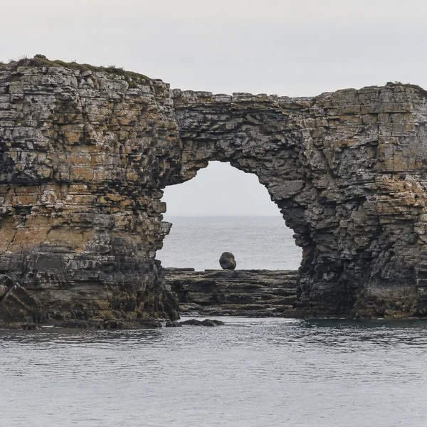 A layered grey and ochre sandstone sea arch frames calm silvery water, with a small dark rounded boulder on the rock shelf in the opening beneath a pale…