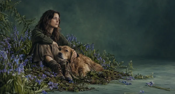 A woman in muted clothing sits among purple-blue flowers beside a golden-brown dog, with a broad blue-green background and open space to the right.
