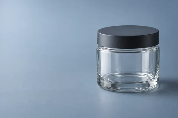 An empty clear cylindrical glass jar with a thick base and matte charcoal screw lid stands on a muted blue-grey surface, positioned on the right with open…