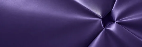 An overhead view of violet satin-like fabric, with glossy angular folds and deep creases gathered across the right side and a broad, relatively flat area on…
