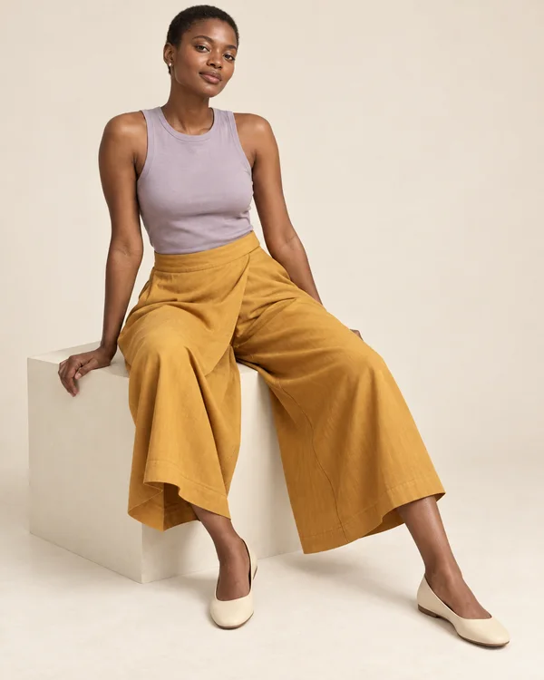 A close-cropped model with deep brown skin sits on an ivory cube against a warm ivory backdrop, wearing a dusty-lilac sleeveless top, saffron wide-leg wrap…