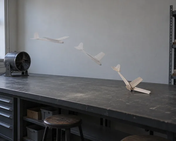 A small pale wooden glider appears in three positions above a charcoal workbench in a grey laboratory: level near the left, descending in the middle, and…
