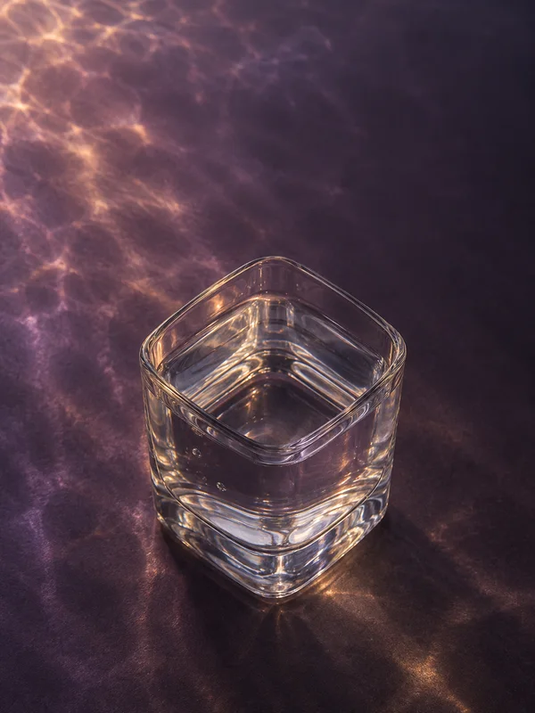 A clear, square tumbler with rounded corners and a thick base sits on a dark plum-colored floor, lit by softly mottled violet and amber highlights; the glass…